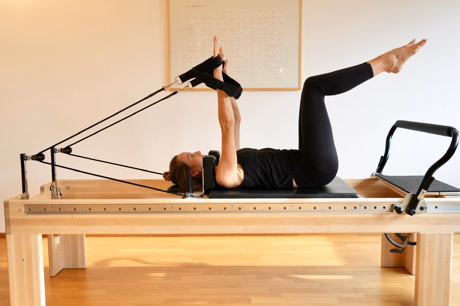 Woman in black workout clothes performing leg and arm exercises on a Pilates reformer machine in a bright room.